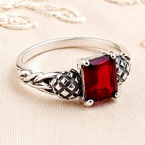 May include: A silver ring featuring a rectangular, deep red gemstone set in the center. The band has a textured, braided metal design on both sides of the stone, with small, grid-patterned metal accents flanking the central red gemstone setting.