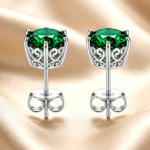 May include: A pair of stud earrings featuring round-cut, deep green gemstones set in silver-toned metal. The settings display an intricate, openwork filigree pattern along the sides, and each earring includes a matching silver-toned post and back.