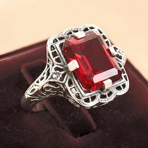 May include: A silver ring with a large, rectangular, deep red gemstone. The ring features an ornate, filigree design around the gemstone and on the band. The setting is square with small silver accents at each corner of the gemstone.