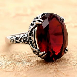 May include: A silver ring featuring a large, oval-cut, deep red gemstone held by four prongs. The band and setting are crafted with intricate, raised floral and vine-like metalwork patterns that extend along the sides of the ring shank.