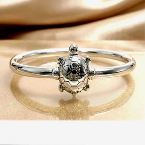 May include: A polished silver bangle bracelet featuring a central turtle charm. The turtle has a detailed, segmented shell design and is integrated into the smooth, rounded band. The metal has a reflective, high-shine finish throughout the piece.
