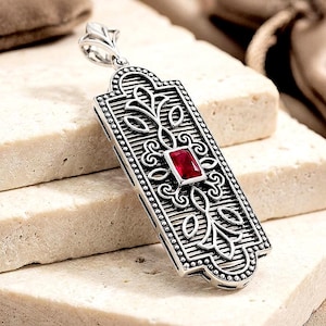 May include: Silver pendant with a rectangular design. The pendant features a central red gemstone and intricate floral patterns. The edges are detailed with small, round accents. The bail has a leaf design.