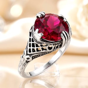 May include: A silver ring featuring a large, round, faceted deep red gemstone held by four prongs. The band and setting display an intricate, dark-oxidized metal lattice pattern with decorative scrollwork details along the sides of the ring.