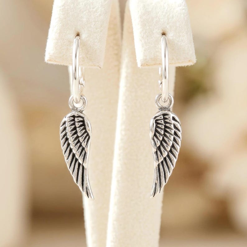 May include: A pair of silver hoop earrings, each featuring a detailed wing charm. The wings have a textured, feathered design, and the earrings are displayed on a white background.