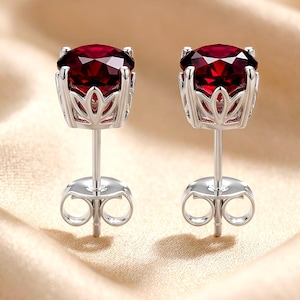 May include: A pair of stud earrings featuring round, deep red faceted gemstones set in silver-toned metal baskets with a petal-like design. Each earring includes a straight post and a matching silver-toned butterfly-style friction back closure.