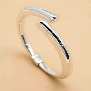 May include: A silver-colored open cuff bracelet. The bracelet has a smooth, polished finish and a minimalist design. The ends of the bracelet are slightly squared off. The bracelet is shown on a neutral background.