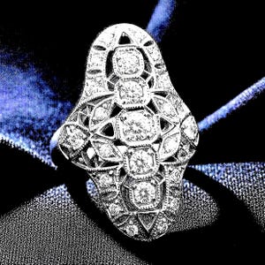May include: A silver-toned elongated ring featuring a vertical row of four round-cut clear stones set within a detailed filigree metal frame. The intricate openwork design is accented with smaller round-cut stones throughout the metal structure.