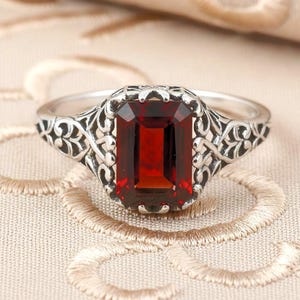 May include: A silver ring featuring a rectangular, deep red gemstone. The gemstone is set in a decorative, filigree-style setting. The ring's band has an ornate, openwork design. The ring is displayed on a neutral-toned surface.