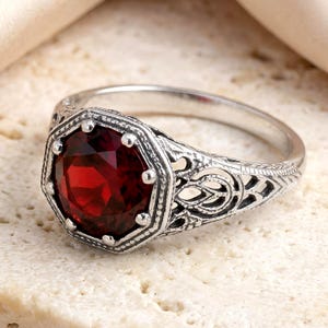 May include: A silver ring featuring a round, deep red faceted gemstone set within an octagonal frame. The band displays intricate, openwork filigree detailing along the sides, with a darkened, oxidized finish highlighting the metal textures.