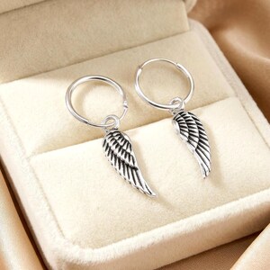 May include: A pair of silver hoop earrings with dangling wing charms. The earrings are displayed inside a beige jewelry box with a satin lining. The wing charms have detailed feather patterns.