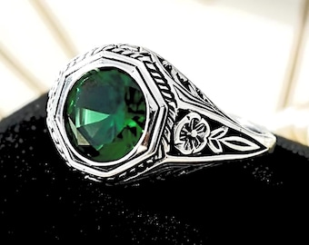 May include: A silver ring featuring a large, round, faceted emerald green stone set within an octagonal frame. The band is decorated with intricate engraved floral patterns and textured detailing along the sides, creating a distinct contrast.