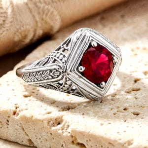 May include: A silver-toned ring featuring a round, deep red gemstone set within a square frame. The band displays intricate, engraved filigree patterns along the sides, showcasing a detailed metalwork design that surrounds the central stone.