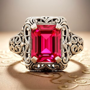 May include: A silver-toned ring with an emerald-cut, vibrant pink gemstone. The ring features an ornate, filigree design around the gemstone and on the band. The gemstone is approximately 0.75 inches long.