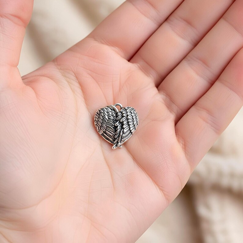 Op de afbeelding: Een kleine, zilverkleurige hartvormige hanger met gedetailleerde vleugelontwerpen. De hanger is ingewikkeld vervaardigd met veervormige texturen en een kleine lus aan de bovenkant voor een ketting. Het hart staat centraal in de afbeelding.