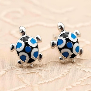 May include: A pair of silver-toned stud earrings shaped like turtles. Each turtle features a black shell with a central blue dot, surrounded by alternating white and blue crescent-shaped enamel patterns on the shell and limbs.