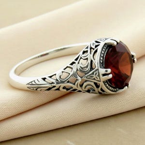 May include: A silver ring with an oval, deep red gemstone. The ring features an ornate, filigree design on the band and around the gemstone setting. The gemstone is the focal point, catching the light and adding a touch of elegance to the jewelry.