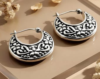 May include: A pair of silver-colored hoop earrings. Each earring features a crescent shape with an ornate floral and swirl design. The earrings are displayed on a light brown surface, with dried flower petals scattered around.