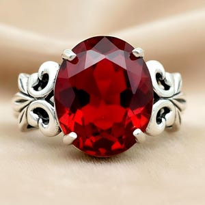 May include: A silver ring featuring a large, faceted, oval-cut red gemstone. The ring has an ornate, swirling design on the band. The gemstone is the focal point, showcasing a deep, vibrant red color.