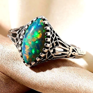 May include: A marquise-cut dark opal ring featuring a vibrant play of green and orange color flashes. The stone is set in a detailed silver band with an intricate, openwork filigree pattern that extends along the sides of the ring setting.