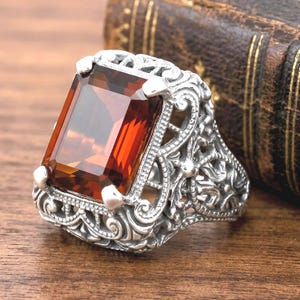 May include: A silver ring featuring a large, rectangular, faceted amber-colored gemstone set in a prong setting. The band and the setting are crafted with intricate, raised scrollwork and filigree metal patterns throughout the entire surface.