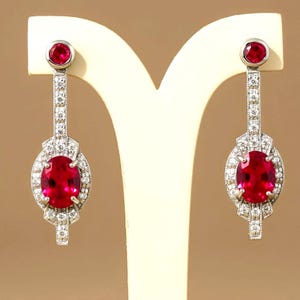 May include: A pair of drop earrings featuring oval-cut vibrant red gemstones set in silver-toned metal. Each earring has a smaller round red stone at the top, connected by a vertical bar encrusted with small clear stones to a larger oval center.