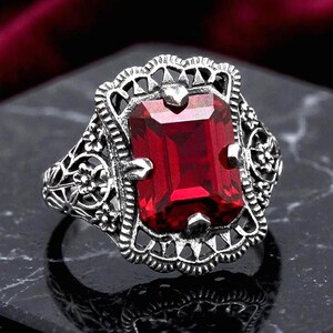 May include: A silver-toned ring featuring a large, rectangular, deep red faceted gemstone set in a decorative, openwork metal frame. The band and setting are adorned with intricate floral and scrollwork patterns, creating a detailed, textured appearance.