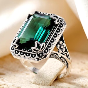 May include: A silver ring featuring a large, rectangular, emerald-green faceted gemstone set in a decorative frame with scalloped edges. The band is detailed with engraved floral motifs and dark oxidized accents, highlighting the intricate metalwork.