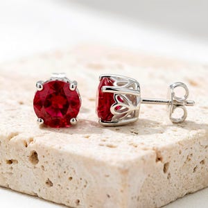 May include: A pair of stud earrings featuring round, faceted, deep red stones set in silver-toned metal. The settings have a decorative, open-work heart pattern along the sides, and the earrings include standard silver-toned metal post backs.