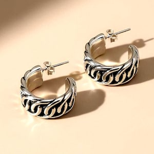 May include: A pair of silver-toned hoop earrings. Each earring features a detailed, textured design along the outer edge, with a series of interconnected, curved shapes. The earrings have a post-and-clutch closure.