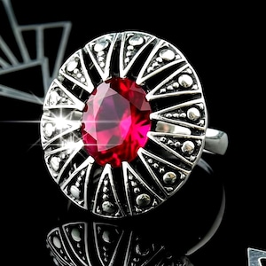 May include: A silver ring featuring a large, oval-cut, ruby-red gemstone. The ring has a detailed, circular setting with triangular and dot patterns. The design is set against a black background, highlighting the ring's intricate details.