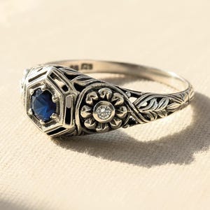 May include: A silver ring featuring a central hexagonal setting with a round blue sapphire, flanked by a circular floral motif containing a small clear diamond. The band displays intricate engraved leaf patterns along the sides and a 925 stamp.
