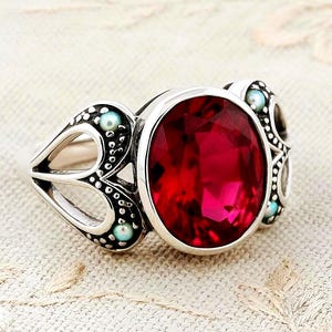May include: A silver ring featuring a large, oval-cut, deep red gemstone at the center. The band is decorated with intricate metalwork, including small, round, light blue accents embedded within the silver frame surrounding the central stone.