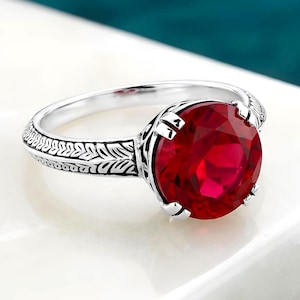 May include: A silver ring featuring a large, round, faceted deep red gemstone set in a prong setting. The band is crafted from silver and displays an intricate, engraved leaf-like pattern along its entire outer surface, extending to the setting.