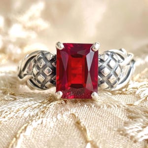 May include: A silver ring featuring a large, rectangular, deep red gemstone. The ring's band has an ornate design with textured details. The gemstone is the focal point, catching the light and showcasing its rich color. The ring is set against a soft, patterned background.