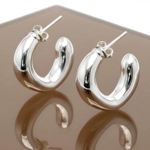 May include: A pair of shiny, silver-colored hoop earrings. The earrings have a smooth, rounded design and a post closure. The earrings are displayed on a reflective surface, which mirrors their shape. The earrings are a classic jewelry item.