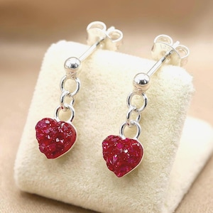 May include: Silver dangle earrings featuring heart-shaped, deep red, crystal-like pendants. The earrings have a post and ball design, with the heart pendants connected by small silver rings. The earrings are displayed on a cream-colored surface.