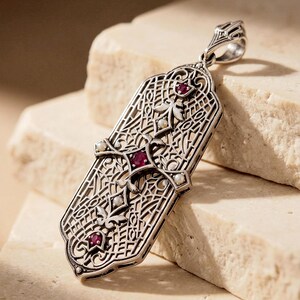 May include: A silver pendant with an intricate filigree design. The pendant features three red gemstones and several small, white pearls. The rectangular pendant has a decorative bail at the top for a chain. The pendant is displayed on a light-colored, textured surface.