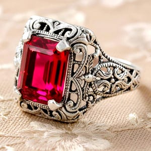 May include: A silver ring featuring a large, rectangular, vibrant red gemstone set in a detailed, openwork metal frame with intricate scrollwork patterns surrounding the stone and extending along the sides of the band.