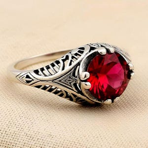 May include: A silver ring featuring a large, round, faceted deep red gemstone set in a prong setting. The band displays an intricate, openwork filigree pattern that extends from the sides toward the central stone, creating a detailed metal design.