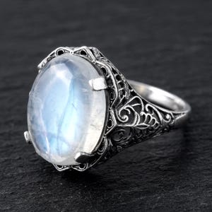 May include: A silver ring featuring a large, oval-shaped, translucent moonstone cabochon with a blue sheen. The metal band displays intricate, openwork filigree detailing along the sides, providing a textured and ornate appearance to the setting.