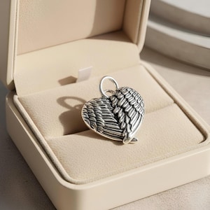 Op de afbeelding: Een zilverkleurige hartvormige hanger met vleugeldetails, gepresenteerd in een beige sieradendoosje. De hanger heeft een lus aan de bovenkant voor een ketting. De vleugels zijn ingewikkeld ontworpen en geven het stuk een delicate touch.