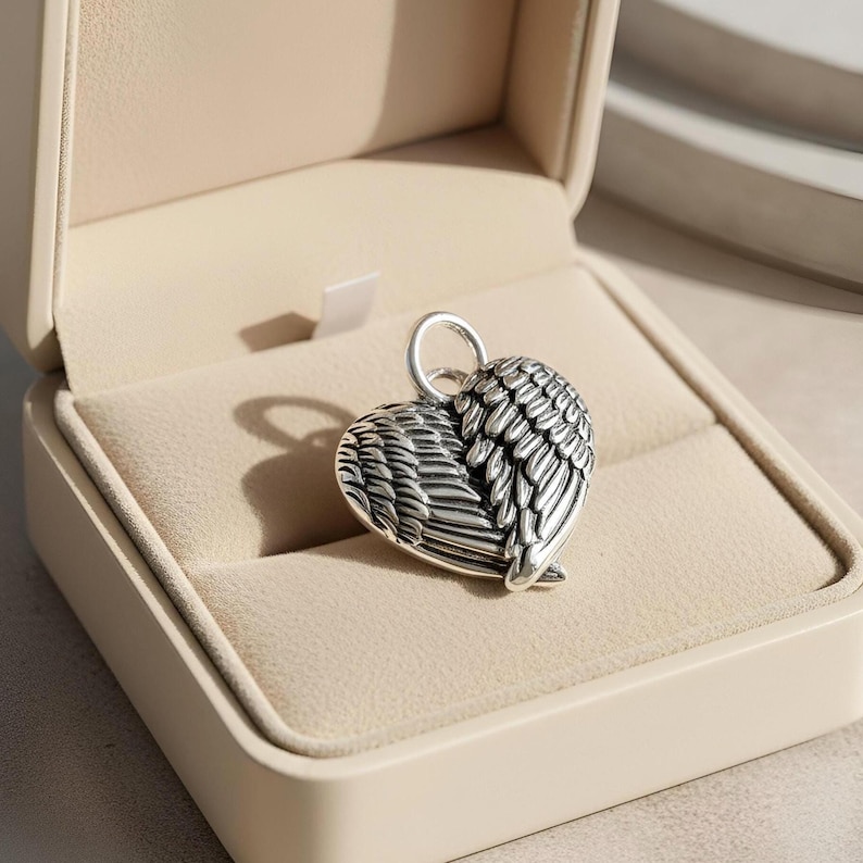 Op de afbeelding: Een zilverkleurige hartvormige hanger met vleugeldetails, gepresenteerd in een beige sieradendoosje. De hanger heeft een lus aan de bovenkant voor een ketting. De vleugels zijn ingewikkeld ontworpen en geven het stuk een delicate touch.