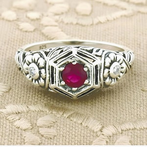May include: A sterling silver ring with a 925 hallmark, featuring a central round-cut vibrant red gemstone set within a hexagonal frame. The band is decorated with embossed floral patterns and two small clear accent stones on either side.