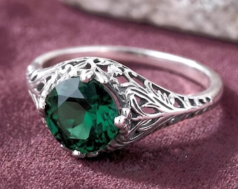 May include: A silver ring featuring a round, faceted dark green gemstone set in a band with intricate, openwork metal filigree detailing that extends along the sides of the setting, creating a detailed, textured appearance around the stone.