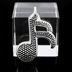 May include: A silver-colored musical note pendant with a textured surface of small, raised dots. The pendant has a loop at the top for a chain and is displayed against a clear, square block on a black background.