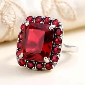 May include: A silver ring featuring a large, square-cut, deep red gemstone surrounded by smaller, round red stones. The ring has a classic design with a polished finish, showcasing the vibrant color of the gemstones.