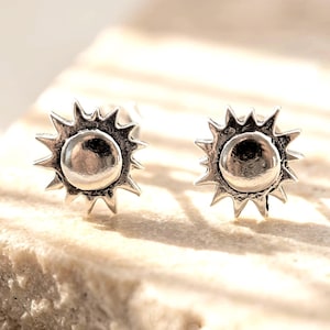 May include: A pair of silver-toned stud earrings featuring a sunburst design. Each earring has a smooth, raised circular center surrounded by pointed, radiating rays. The metal has an oxidized finish in the recessed areas to provide contrast.