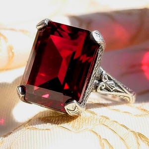 May include: A ring featuring a large, rectangular, deep red gemstone set in a silver-toned metal band. The metal band displays intricate, raised decorative patterns along the sides, and the stone is secured by four prominent corner prongs.