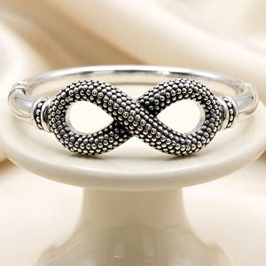 May include: A silver-toned bracelet featuring an infinity symbol design. The infinity symbol is textured with small, raised dots, creating a detailed pattern. The bracelet is displayed on a white pedestal.