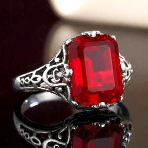 May include: A silver-toned ring featuring a large, rectangular, faceted red gemstone set in a decorative, openwork metal band with intricate scroll-like patterns. The vibrant red stone is the central focus, held by small prongs.